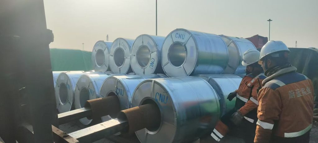 Workers and coil-handling equipment beside wrapped coils marked CNU.