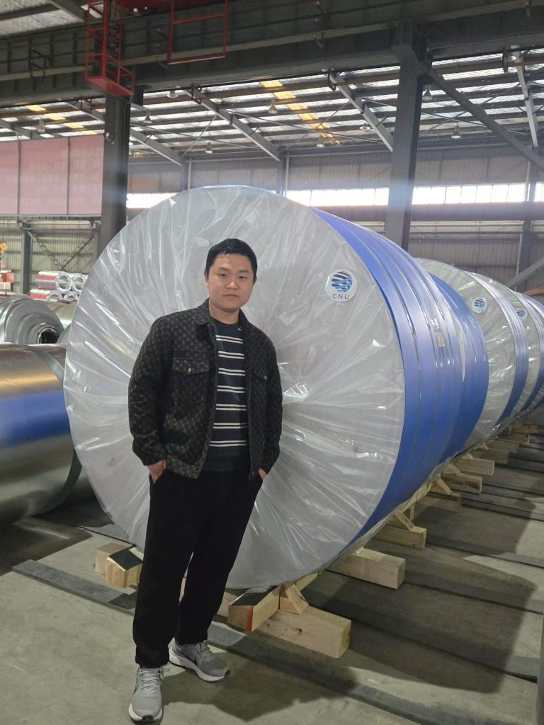 A wrapped coil bearing a CNU label, supported on timber in the factory.