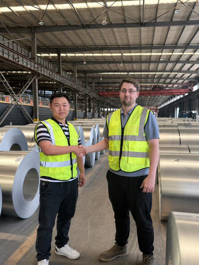 Factory visitors wearing reflective vests beside rows of metal coils.